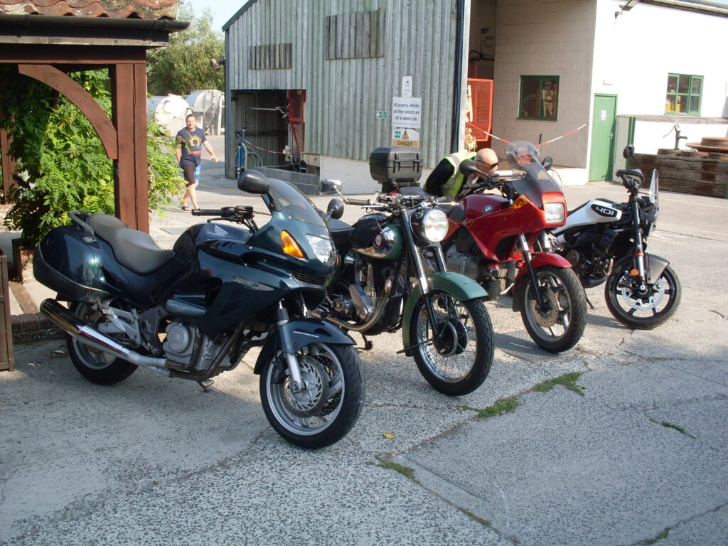 Four very different motorcycles parked in a line. One quite modern, dark green, with a fairing and screen; next to it an older lighter-green unfaired machine; then a red machine with an angular fairing and screen and lastly a smaller, almost new machine with a handlebar-mounted screen and a white-panelled one-piece tank and seat. Behind are two buildings, one very agricultural and one showing the end of a low. tiled, brown wooden veranda .