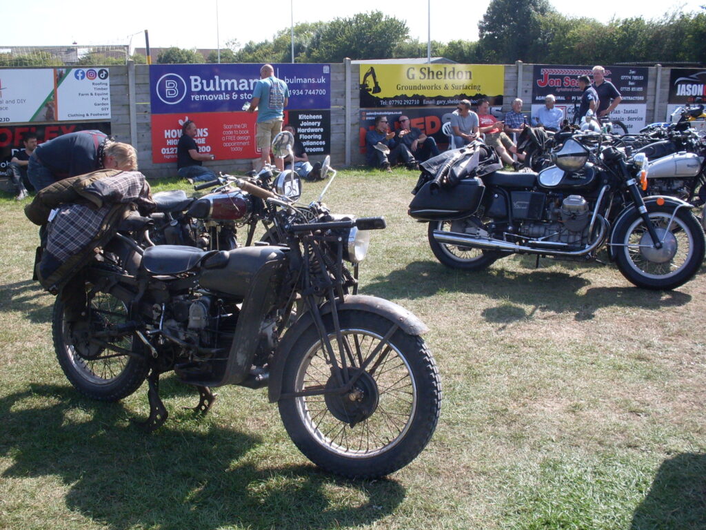 Several motorcycles parked on brown-green grass in front of several people relaxing against a fence of advertising hoardings. The nearest machine is khaki-brown with old girder-type forks, legshields, a single saddle and what looks like a big coat piled over the back. Slightly apart from it is a much more modern machine with a chromed exhaust pipe and silencer running along one side, a helmet slung from the handlebars, black panniers over the rear wheel with a motorcycle jacket slung over.
