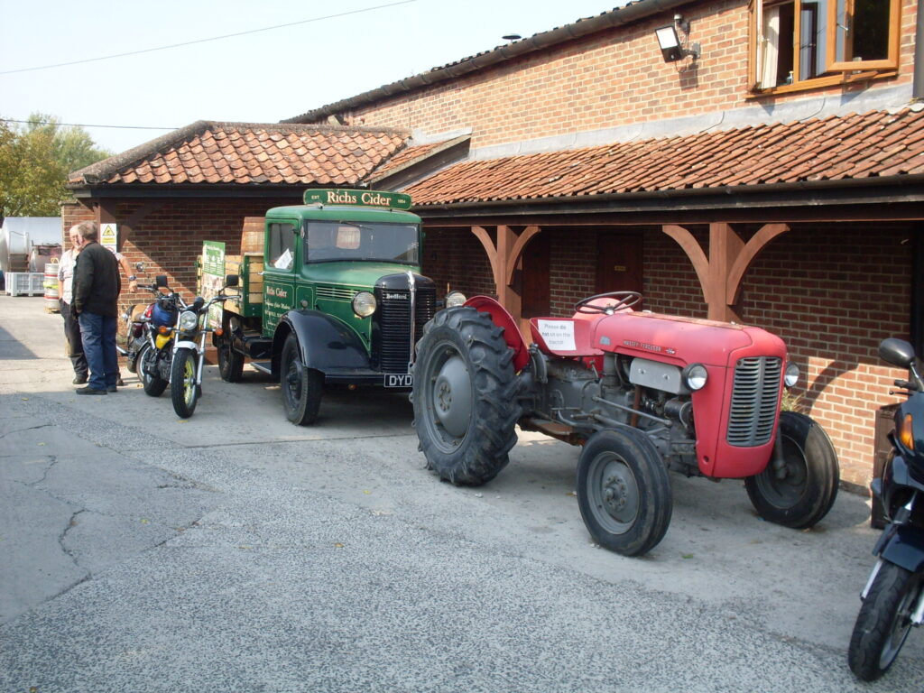 An old red tractor and an old green lorry with a wooden flatbed and a 'Richs Cider' board across the top of the cab are displayed in front of a red-tiled brick building with a low wooden veranda along its side.