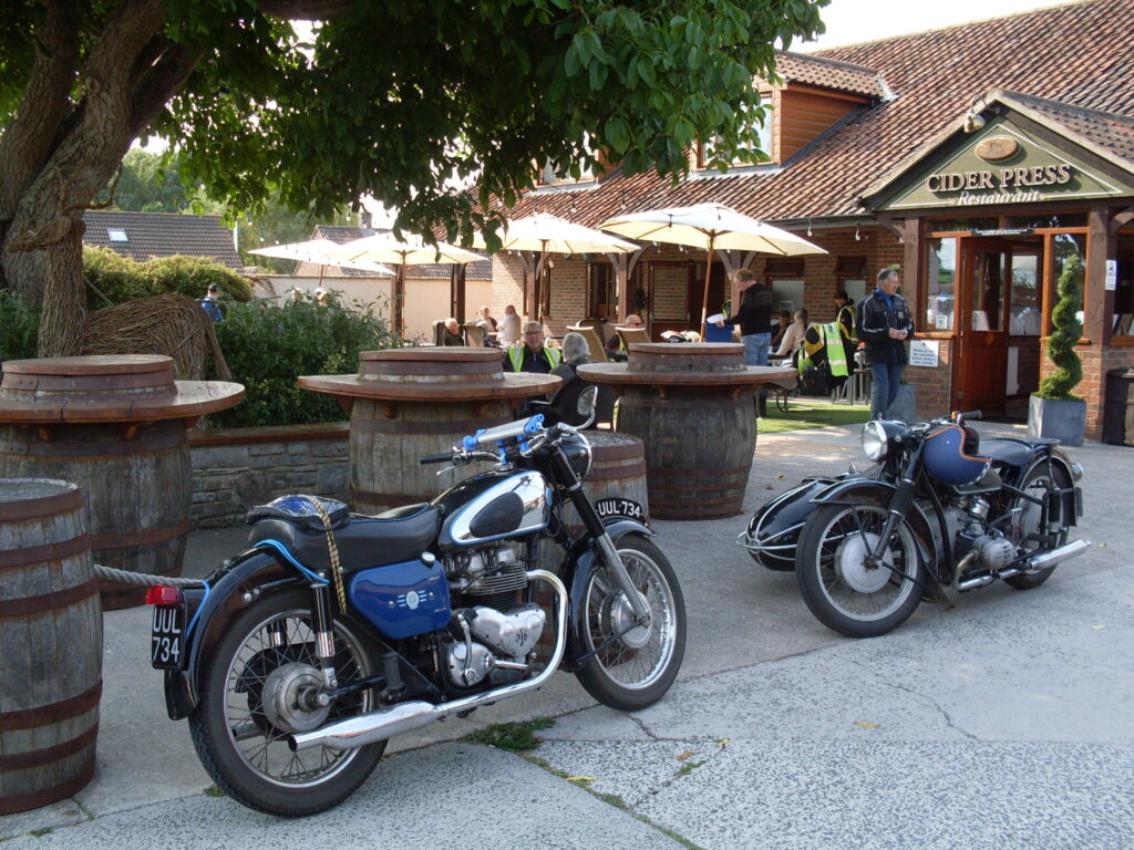 A shiny blue, black and chrome motorcycle parked facing an equally shiny black motorcycle combination. Behind them some big wooden barrels converted into tables and behind again several tables and chairs, under sunshades, with people sitting and standing. In the background a low red-brick restaurant building with a big tiled roof.