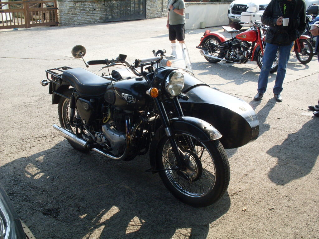 A black BSA 650cc twin motorcycle and sidecar, parked in the sun with two onlookers (the nearest holding a mug of tea or coffee). In the picture both onlookers are cut off at the shoulders.
