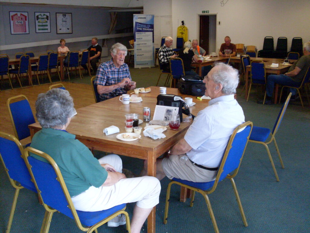 Inside the Clubhouse, three people sit on blue chairs, around a brown wooden table on which can be made out cups of tea, scones on plates and small containers of white cream and red jam. A bit apart behind them are more people, tables and chairs.