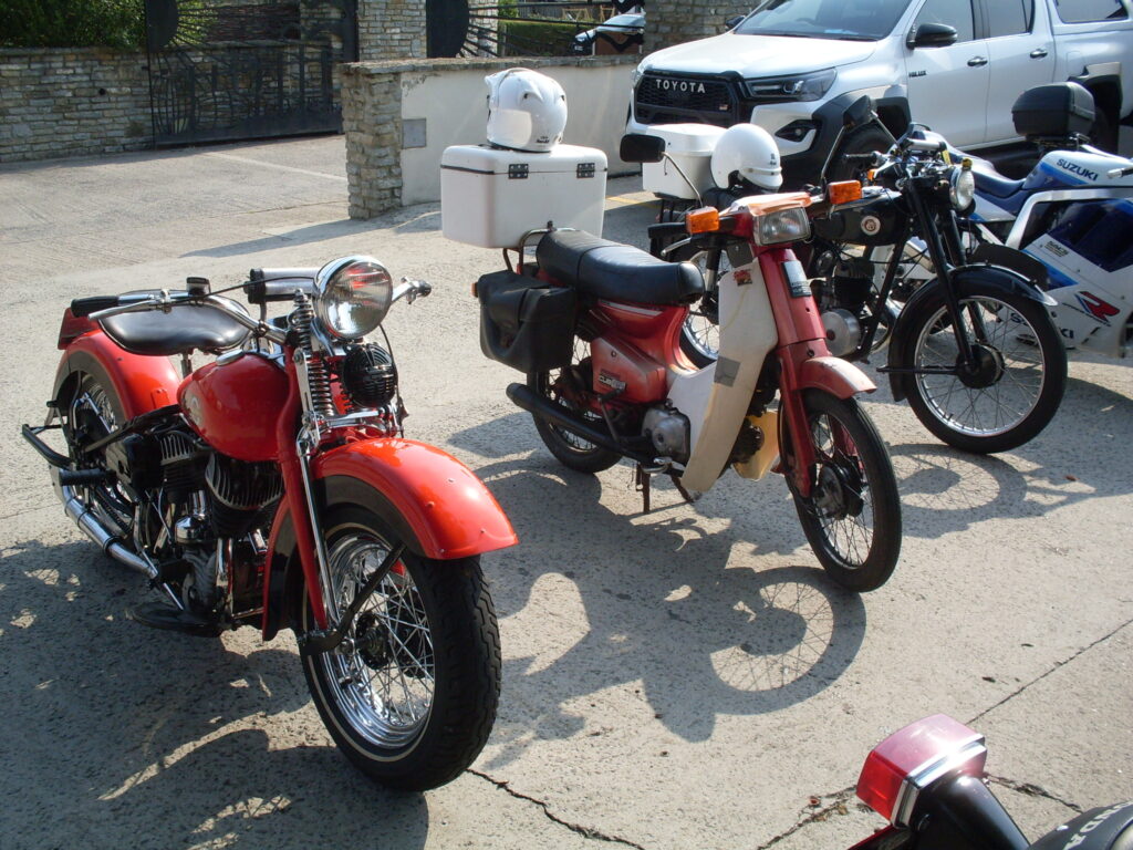 Three parked motorcycles, a red V-twin Harley Davidson, a red and white 90cc step-through Honda and dark-green two-stroke Francis Barnett 'lightweight'.