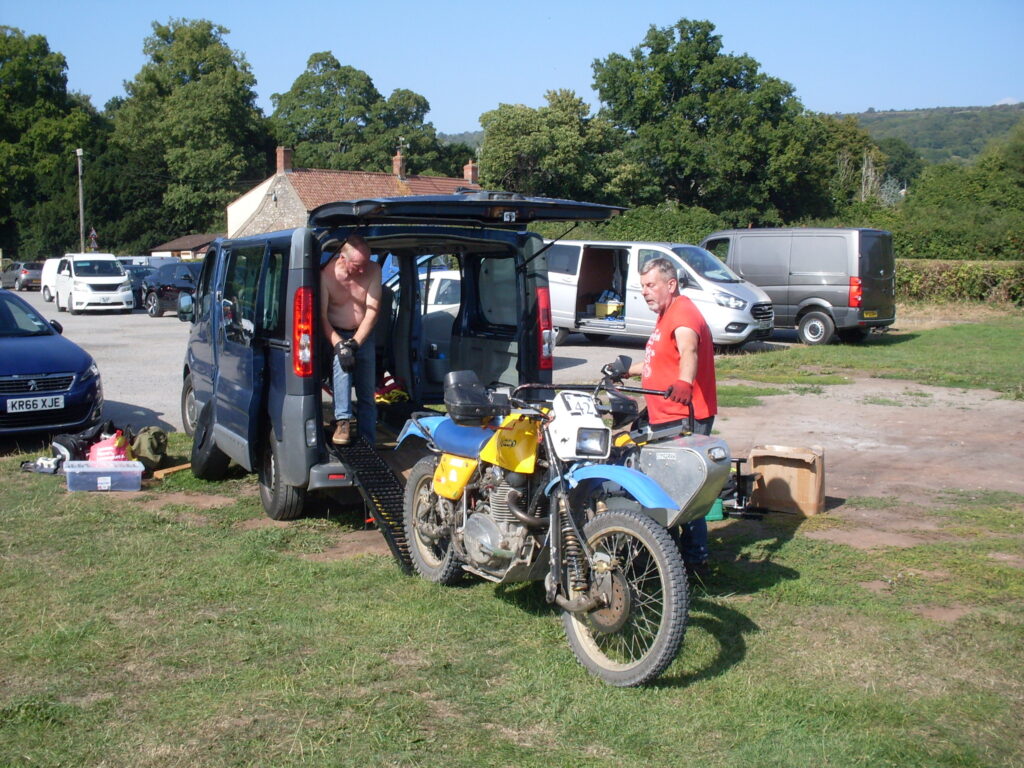 The blue and yellow Wasp outfit rear-on to a dark blue van with the back door raised wide open and a ramp down. A shirtless man in the van doorway and another (in a red T-shirt) behind the outfit look to be discussing exactly how to load things up!