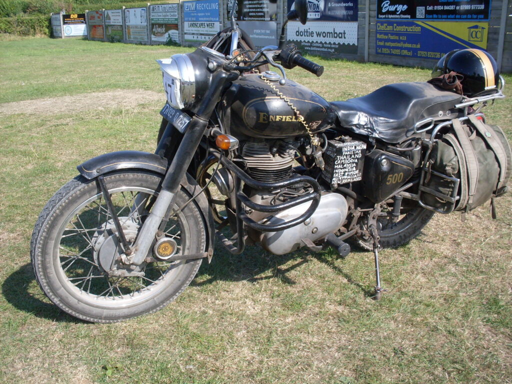 Parked on teh grass, on its own, with a fence of advertisements behind, a black single-cylinder machine. On its rear rack rests a black open-face helmet with a gold strip over its top; one of a pair of canvas pannier bags bulges beside the rear wheel. Behind the engine, under the saddle, the side of the battery-box is covered in a white-printed list of many countries that have been visited.