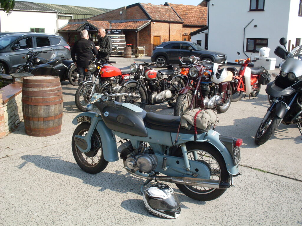 Several parked motorcycles, with a couple of cars and two men chatting behind, and a background of various buildings (two with white walls, one of brick with a red-tiled roof).
