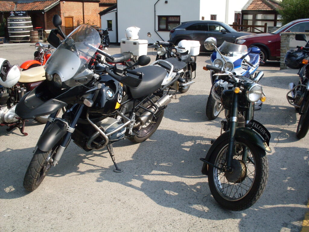 Several parked motorcycles, with cars and buildings (and the occasional big wooden barrel) behind. Prominent in the foreground a big, bulky, black modern BMW almost towers over an old dark green Francis Barnett 'Falcon' lightweight two-stroke.