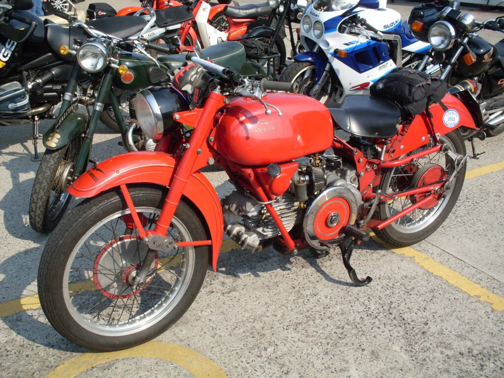 In front of many other parked motorcycles, a bright red Italian Moto Guzzi machine, its engine with a single horizontal cylinder and a big red and chrome disc of a flywheel at the side right next to the rider's footrest.