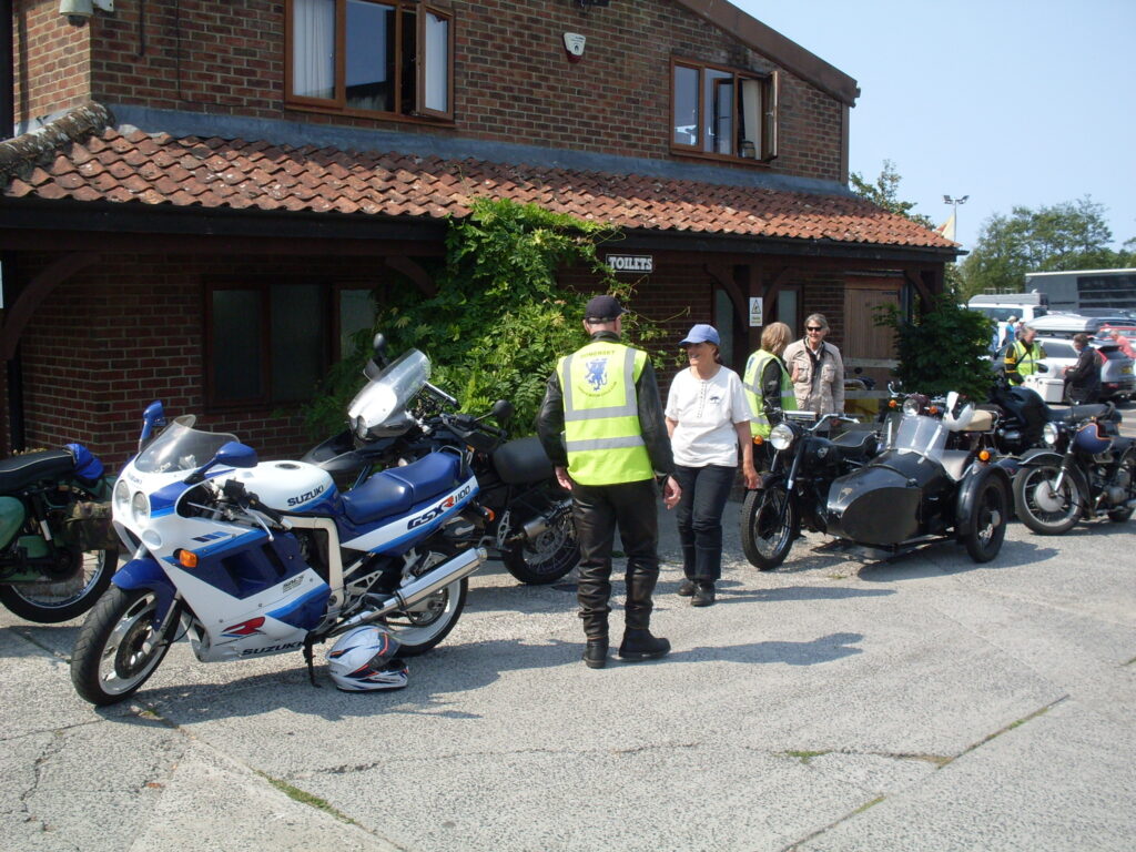 Several motorcycles parked in front of a brick building with a low tiled veranda. Groups of people are standing among the bikes, chatting.