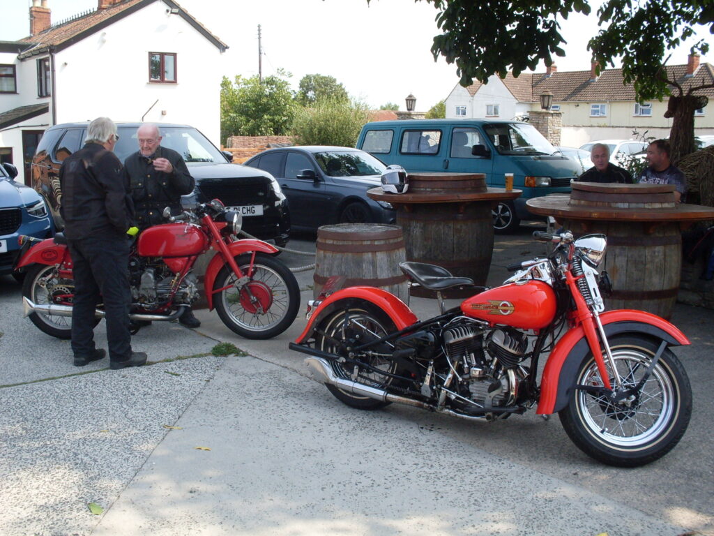 The big red Harley and the red Guzzi parked in front of big wooden barrels (on their ends, acting as tables). Two rider stand either side of the Guzzi, in a discussion.