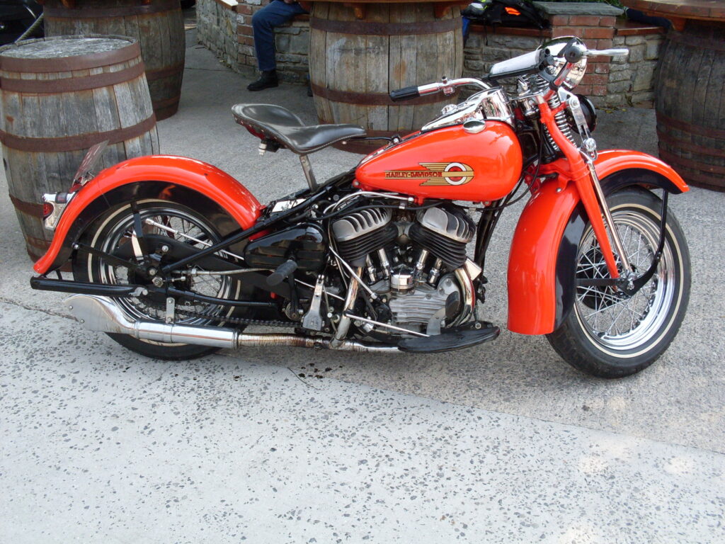 A picture of the red V-twin side-valve Harley Davidson, with its massive single seat saddle sticking up behind the petrol tank, parked in front of several big wooden barrels standing on their ends.