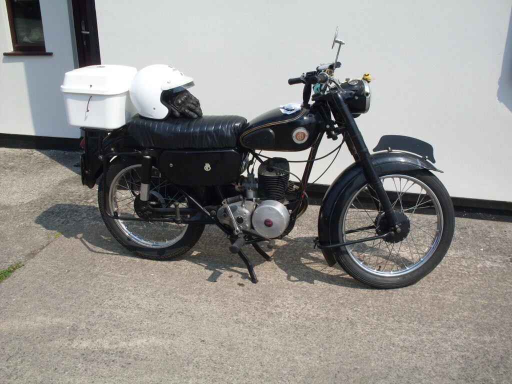 A picture of a small single-cylinder two-stroke lightweight Francis Barnett - almost black-looking against a white wall in bright sunlight. It has a white top-box mounted at the back and a white open face helmet resting on the black dualseat.