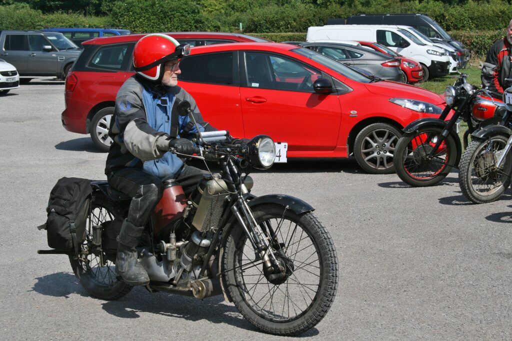 A rider on a motorbike with a crimson horseshoe tank under the riders saddle and black throw-over pannier bags over the rear rack coasts into the car park. He is dressed in black leather riding boots and trousers and a blue and grey jacket. On his head a red crash helmet with white edge trim and black goggles. In the background red, grey and white vehicles and the front wheels of several parked motorbikes show at the right hand side.