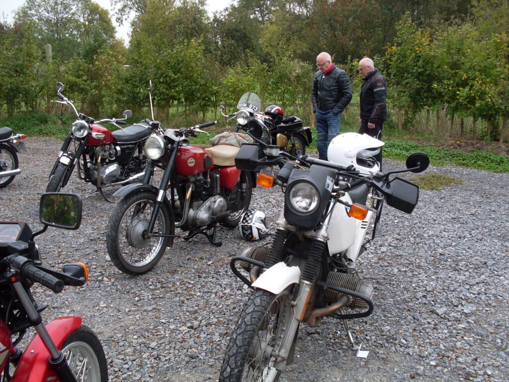 Several motorcycles parked on coarse grey gravel with a background of small trees  and grass.  In front a white BMW, a red Ariel and a red Triumph. Behind these just in view a smaller motorcycle with a flyscreen, racing handlebars and two men in discussion next to it. 