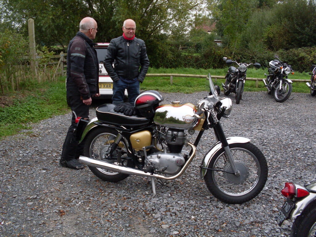 Two men in riding gear standing next to a single-cylinder Royal Enfield from the 1960s with shiny chrome-plated mudguards and petrol tank and a sporty flyscreen and exhaust.