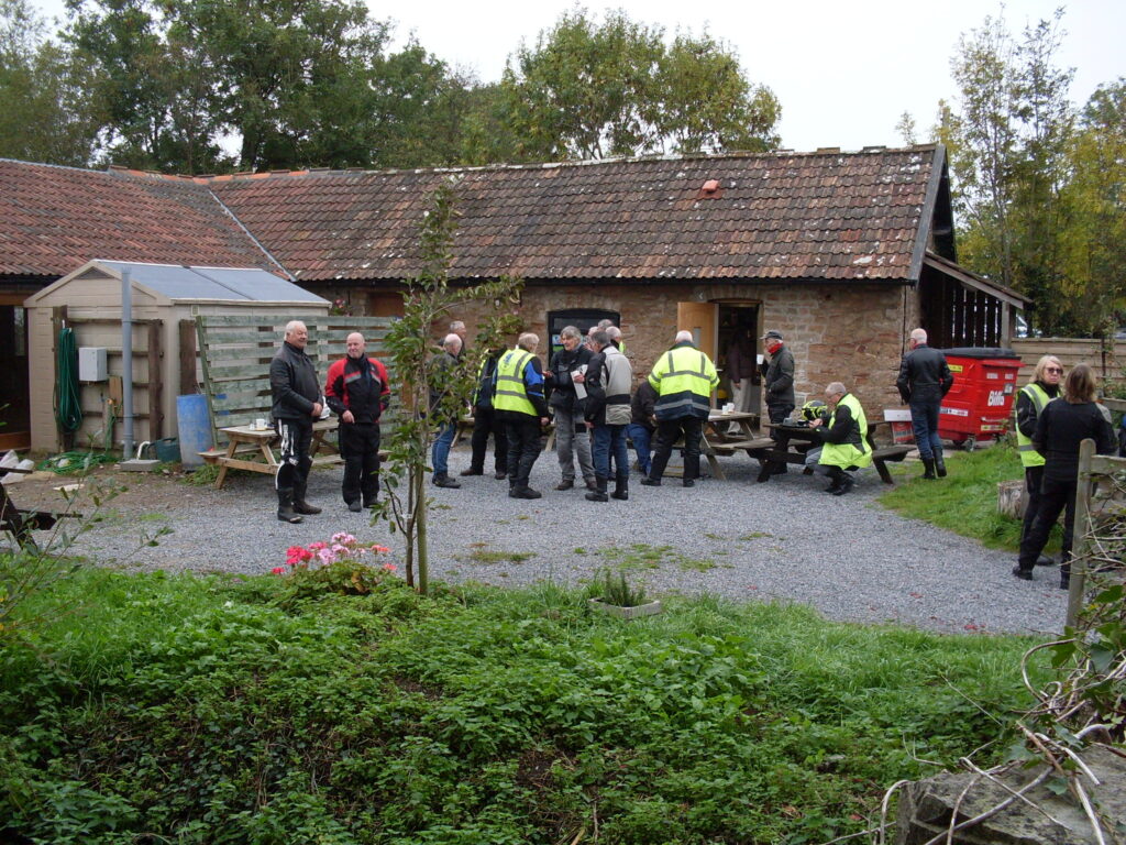 A closer view of the bikers around the tables with green foliage in the foreground and the single-storey tiled-roof building  behind.