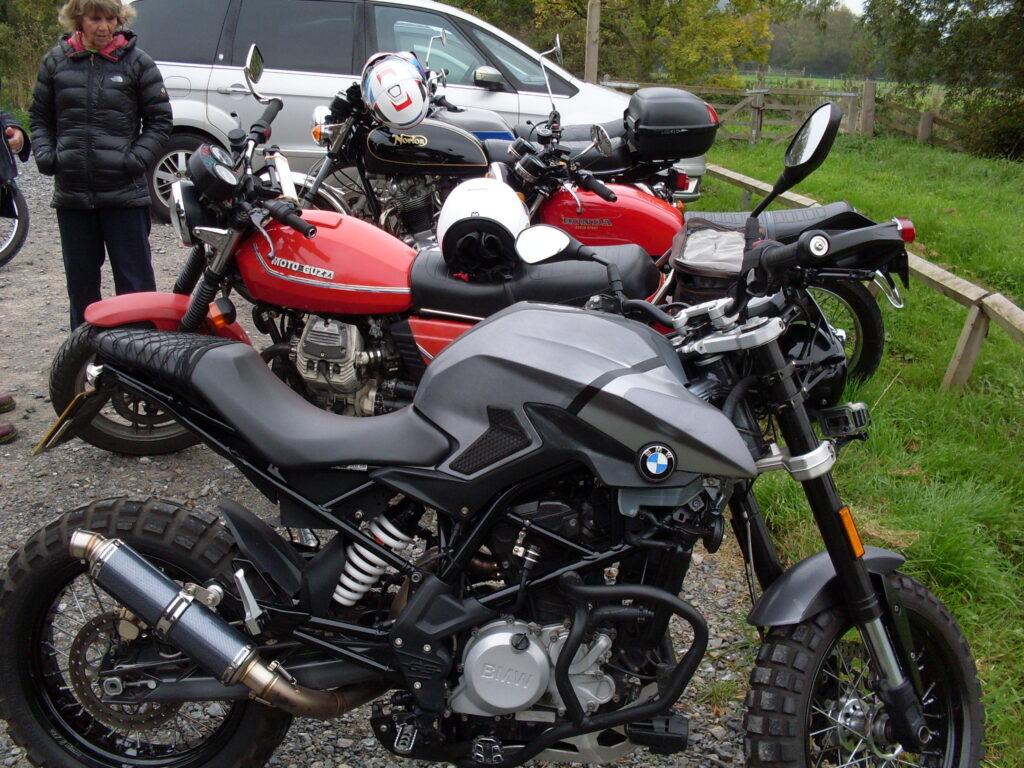 Several motorbikes in line viewed one behind each other. In the foreground a bike with a BMW badged tank, a tangle of pipes that are a mix of frame and exhaust system and a single-cylinder engine that leans backwards.