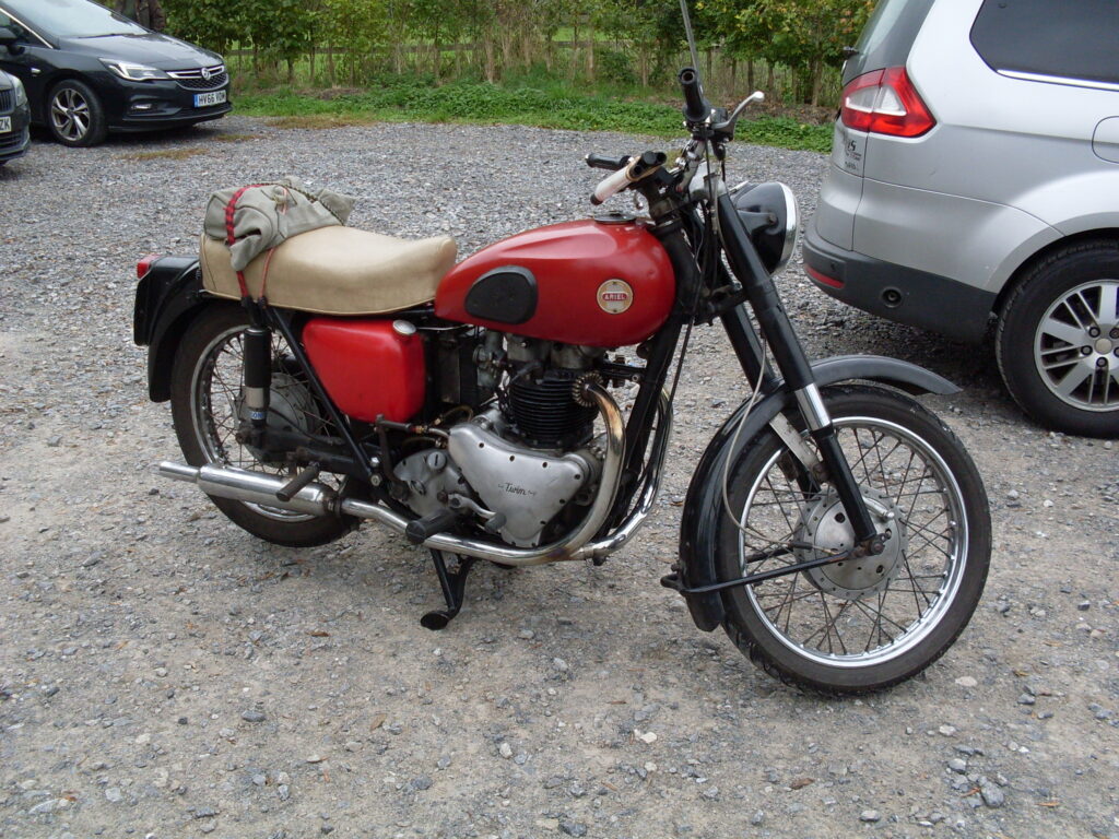 A red and black motorbike on its own, with cars to left and right and small trees and a wooden fence behind.