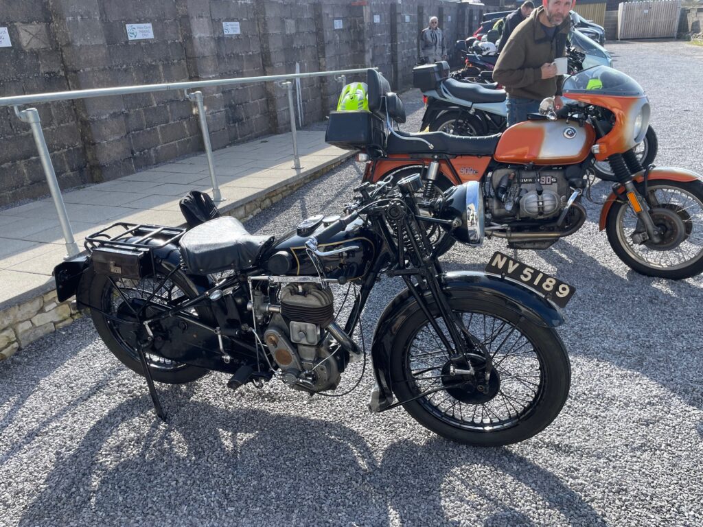 A study of the black 1935 Sunbeam with its big single saddle. Immediately behind is a much more modern BMW  R90S 'boxer' twin from the 1970s with its ornage and silver paintwork and small 'bikini'-style handlebar fairing 