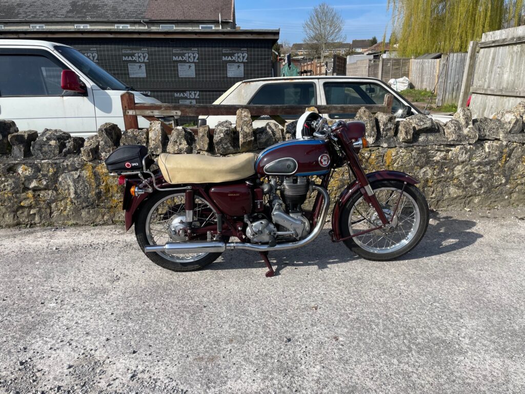 A clean and shiny dark red motorcycle with a beige dualseat is parked on its own in front of a low stone wall.
