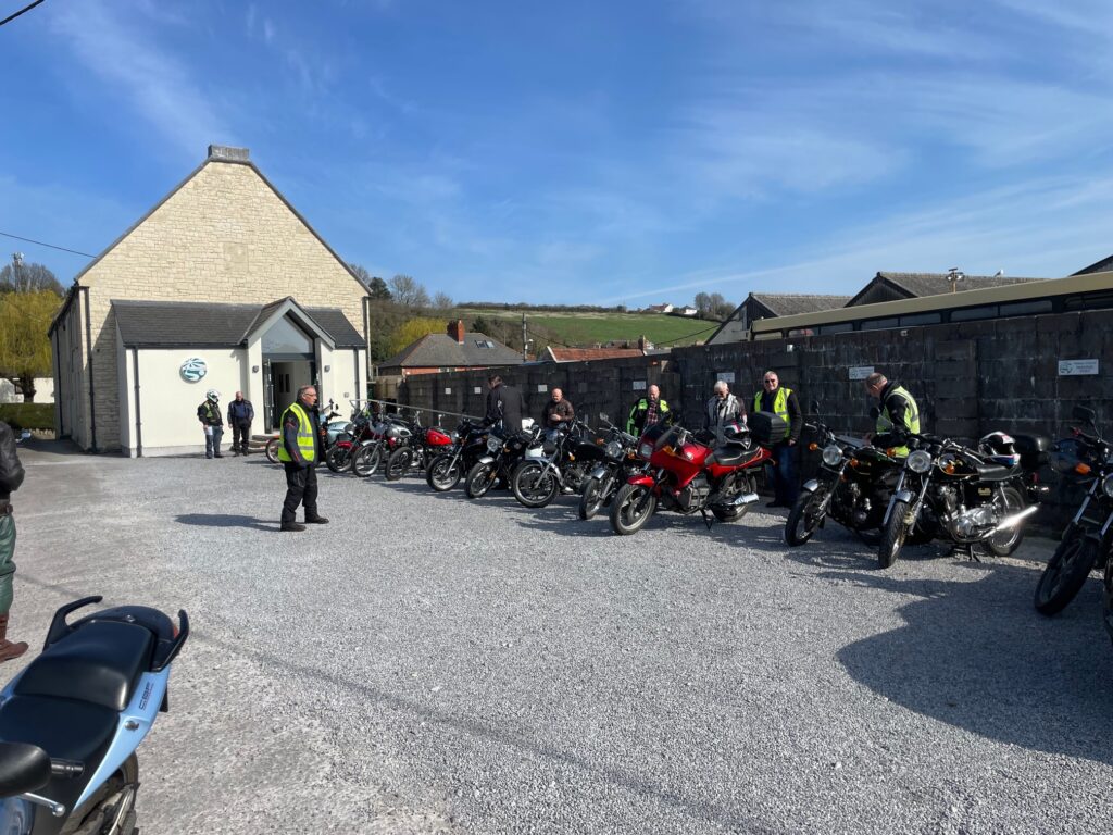 A line of motorcycles parked along a high wall bordering a gravel car-park, under a blue sky. In the distance a cream gable of a two-storey building. Riders move between the motorcycles, viewing and discussing.