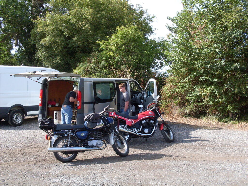 Two motorcycles parked alongside a van which has a man and a boy bending into it.