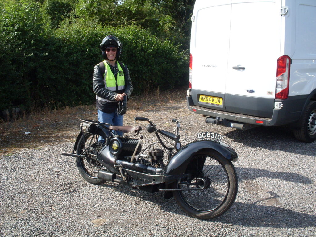 A man with a crash helmet, dark sunglasses and a very big smile stands behind a low black spindly machine (looking like a 1920s motor scooter).