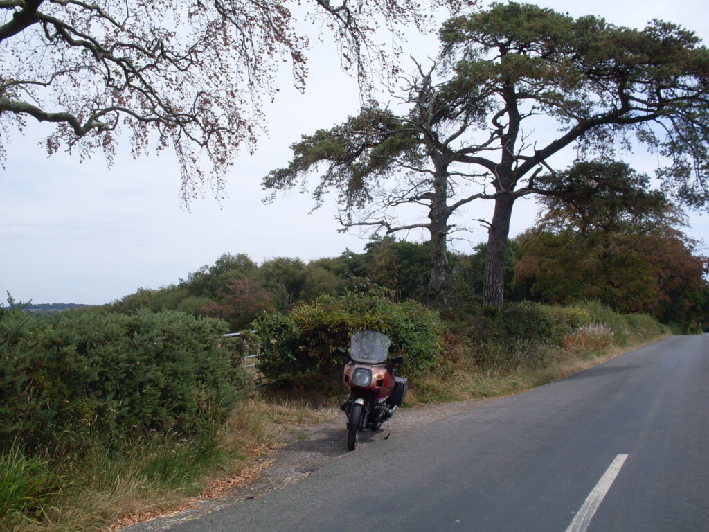The same motorcycle as before parked at the side of the road with a background of trees, hedges and a farm gate