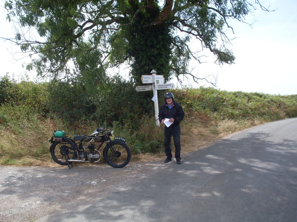 The newly arrived rider has parked his 1920s  machine next to a signpost (under a tree!)  - he has some paper notes in his hands and grins at the camera.